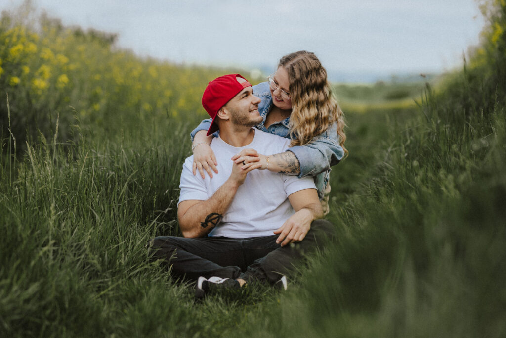 Fotograf Reutlingen Tübingen Hochzeit Paar sitzt an einem Rapsfeld und sie umarmt ihn von hinten in Pliezhausen